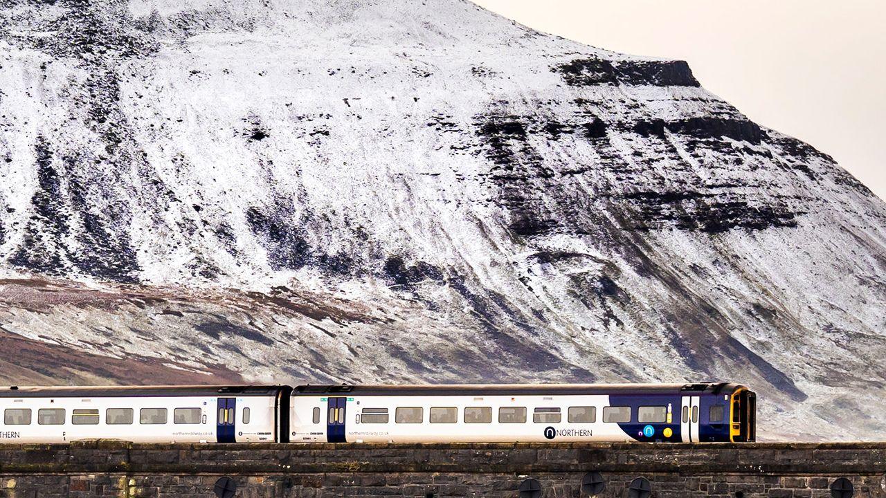 A Northern train travels over a stone bridge with snow on a rocky hill in the background in the Yorkshire Dales