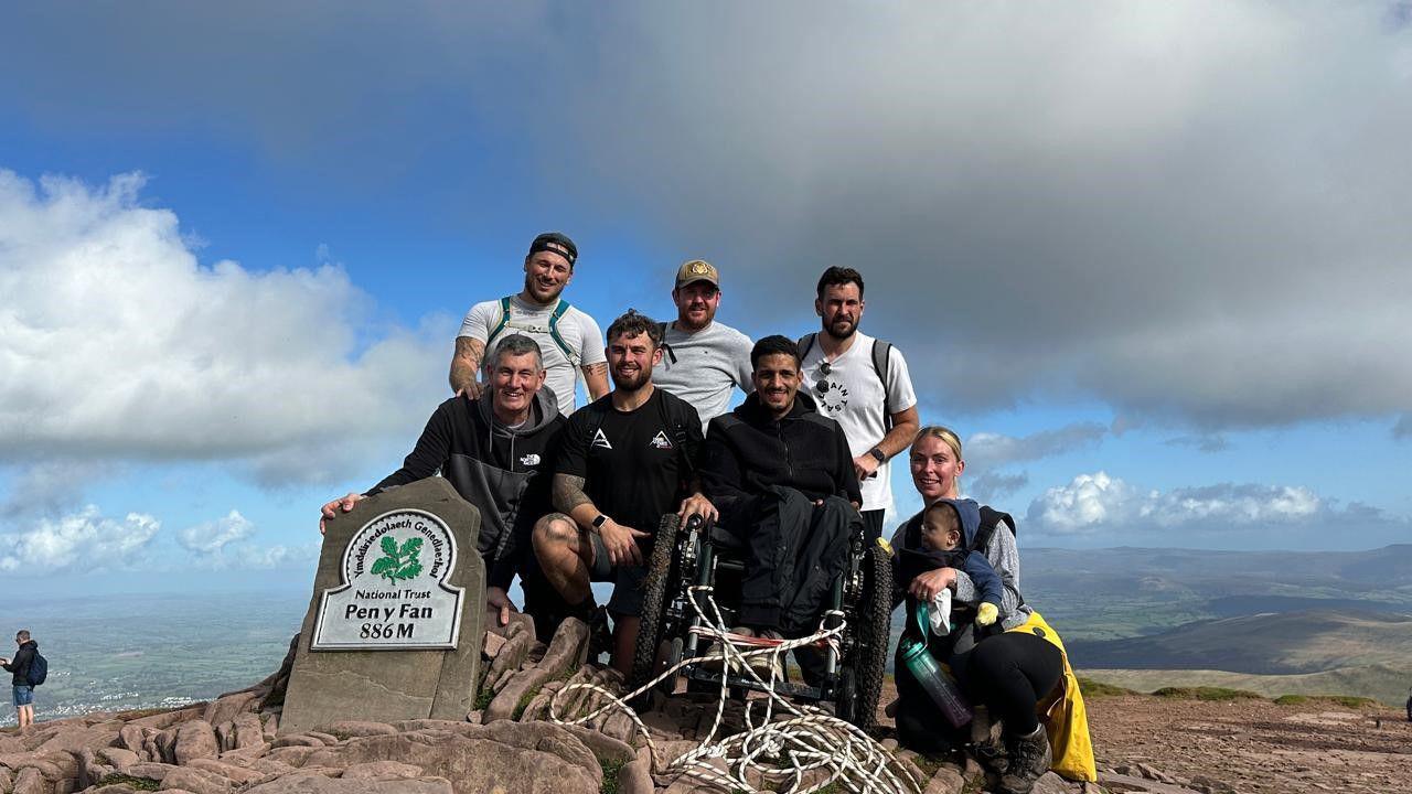 A group of people at the summit of Pen y Fan. Liam is pictured centre right in his Mountain Trike. He is surrounded by five other men. There is a woman holding a child kneeling next to him.