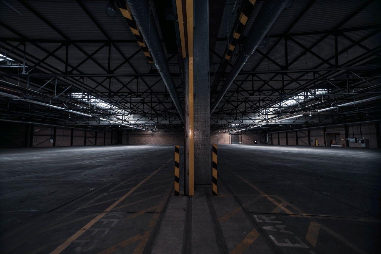 The interior of a large empty warehouse-like building. Steel supports are on the roof and a large support beam with black and yellow tape attached is in the middle of the photo