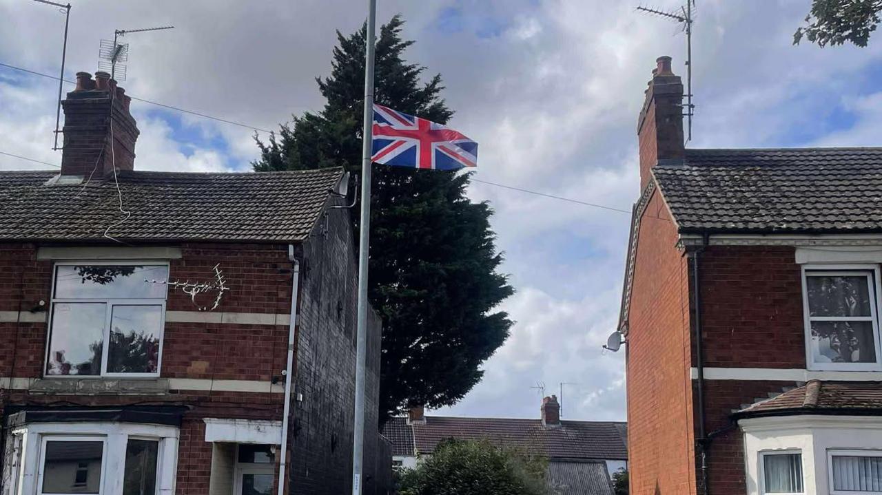 A Union Flag, flying on a lamp post, with two rows of red brick terraces homes either side. There is a large free behind it and a sky full of clouds. 