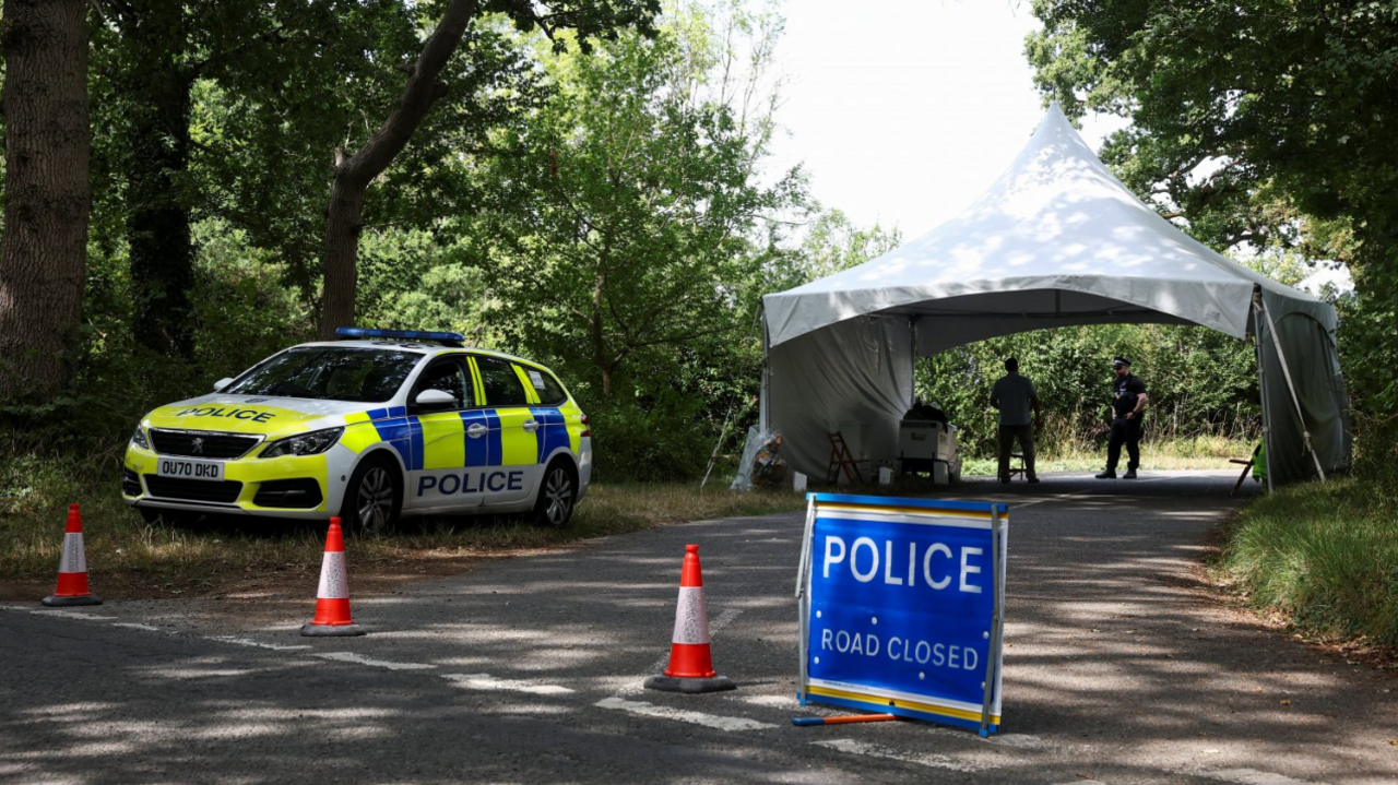 A police car parked by the side of a rural country road with a blue police road closure sign and traffic cones blocking the way. Two police officers stand under a gazebo just behind the block