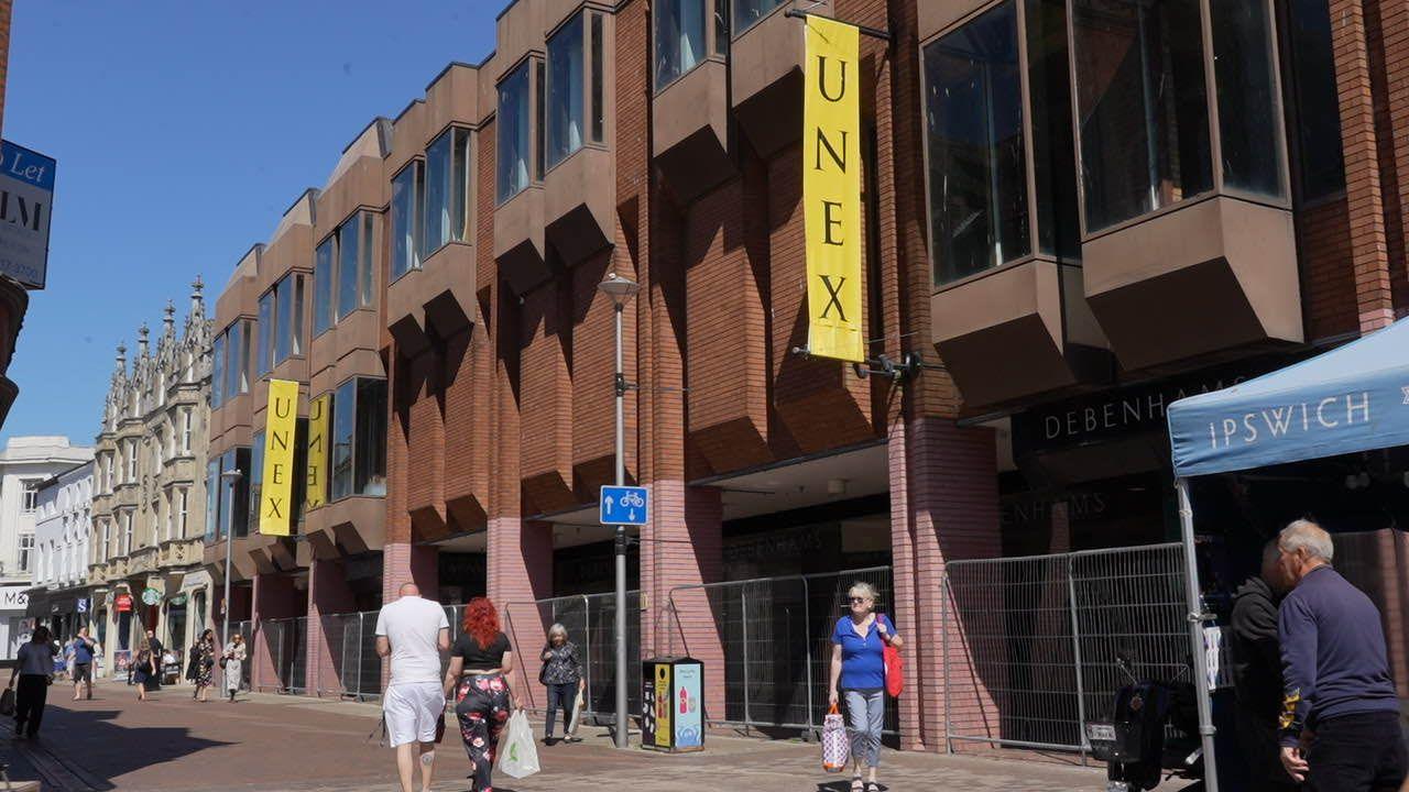 Westgate Street in Ipswich on a sunny day. The former Debenhams is shown with fencing blocking off its perimeter as shoppers go about their business.