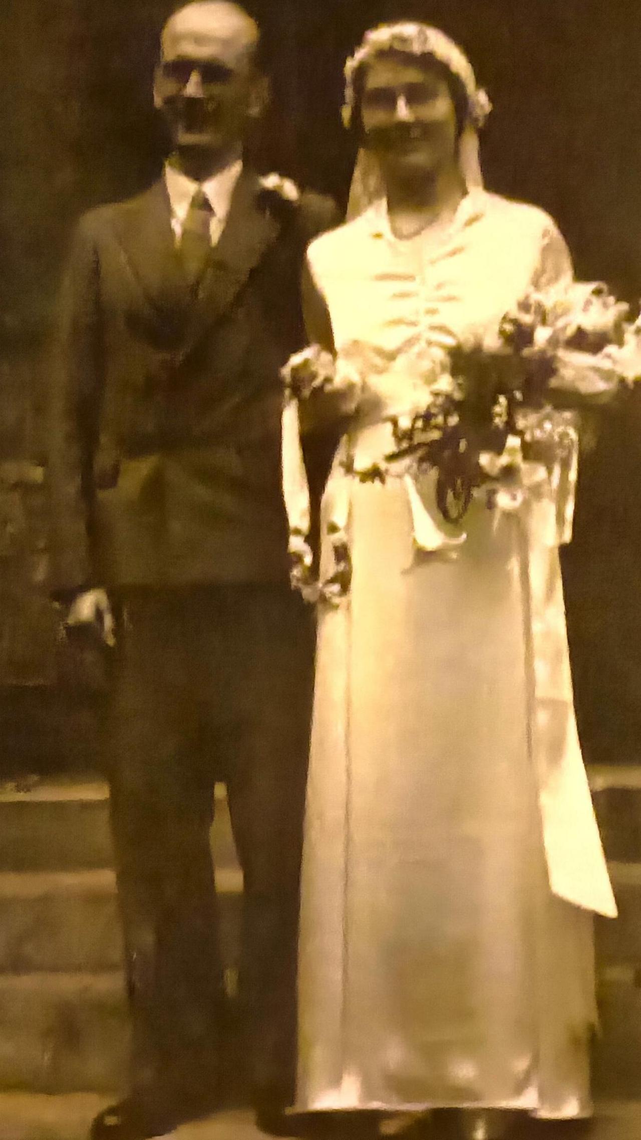 A black and white photo of a couple in wedding clothes linking arms. She is holding a bouquet and wearing a veil. 