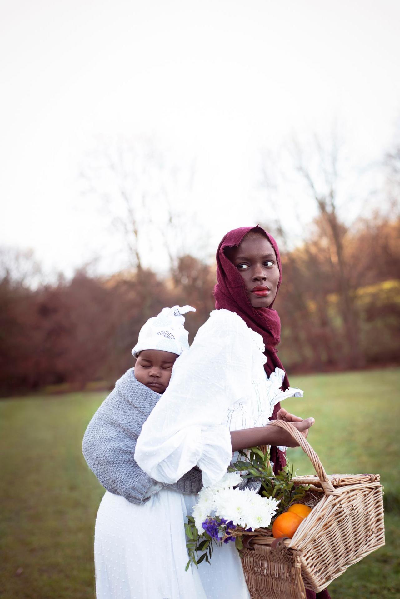 Dola Posh is outside, dressed in a white rob and red headscarf, carrying her baby in a blue blanket on her back. She's carrying a wicker picnic basket.