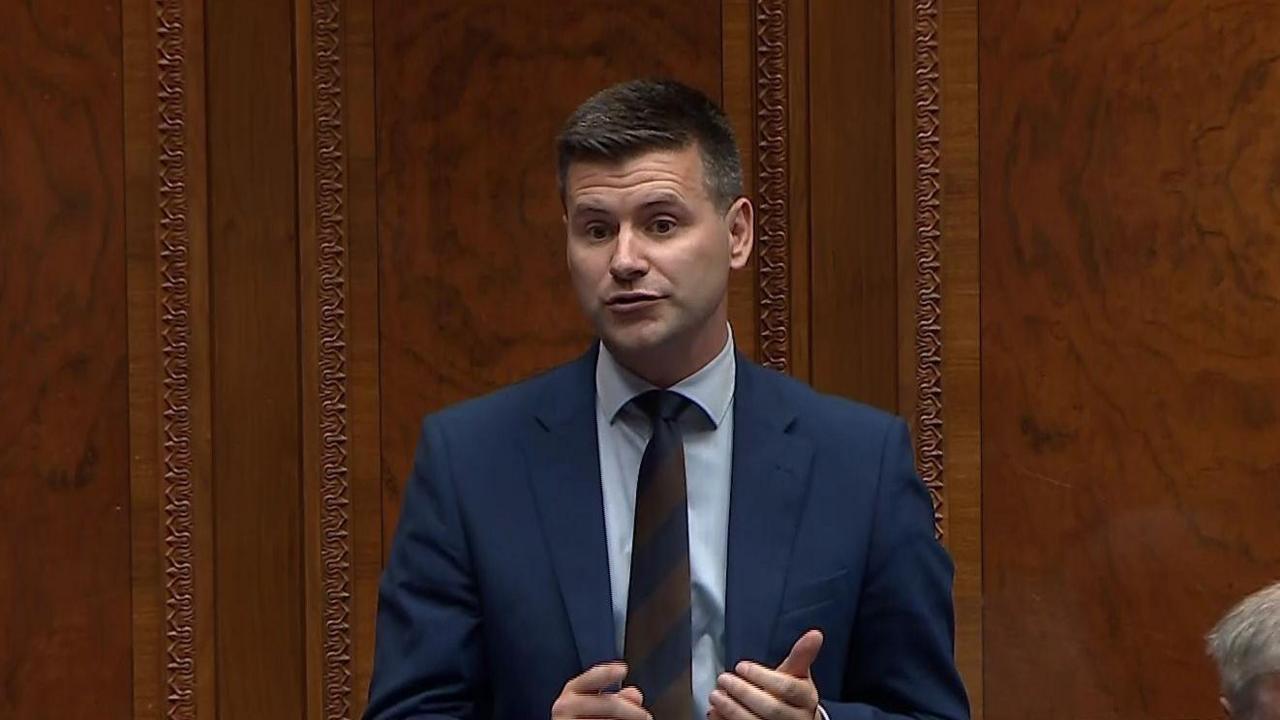 A man stands in front of a brown wood panelled wall. He wears a blue suit, blue shirt and brown and navy striped tie. He has short dark hair.