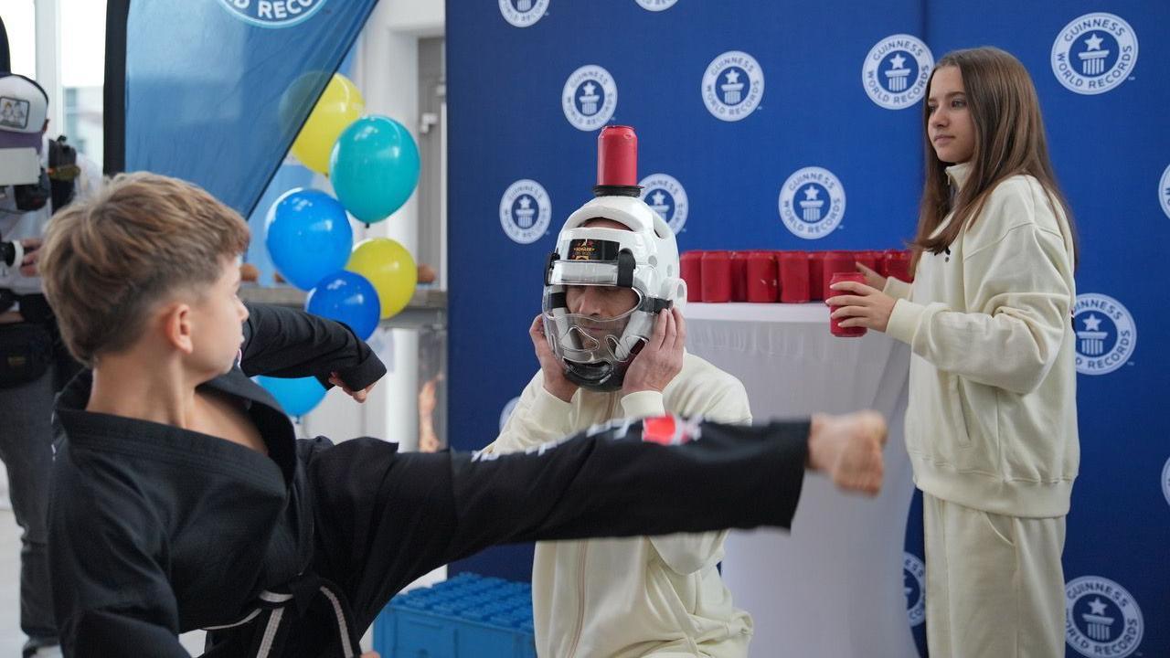 A boy wearing martial arts robes kicks a can off a person's head, who is wearing a helmet