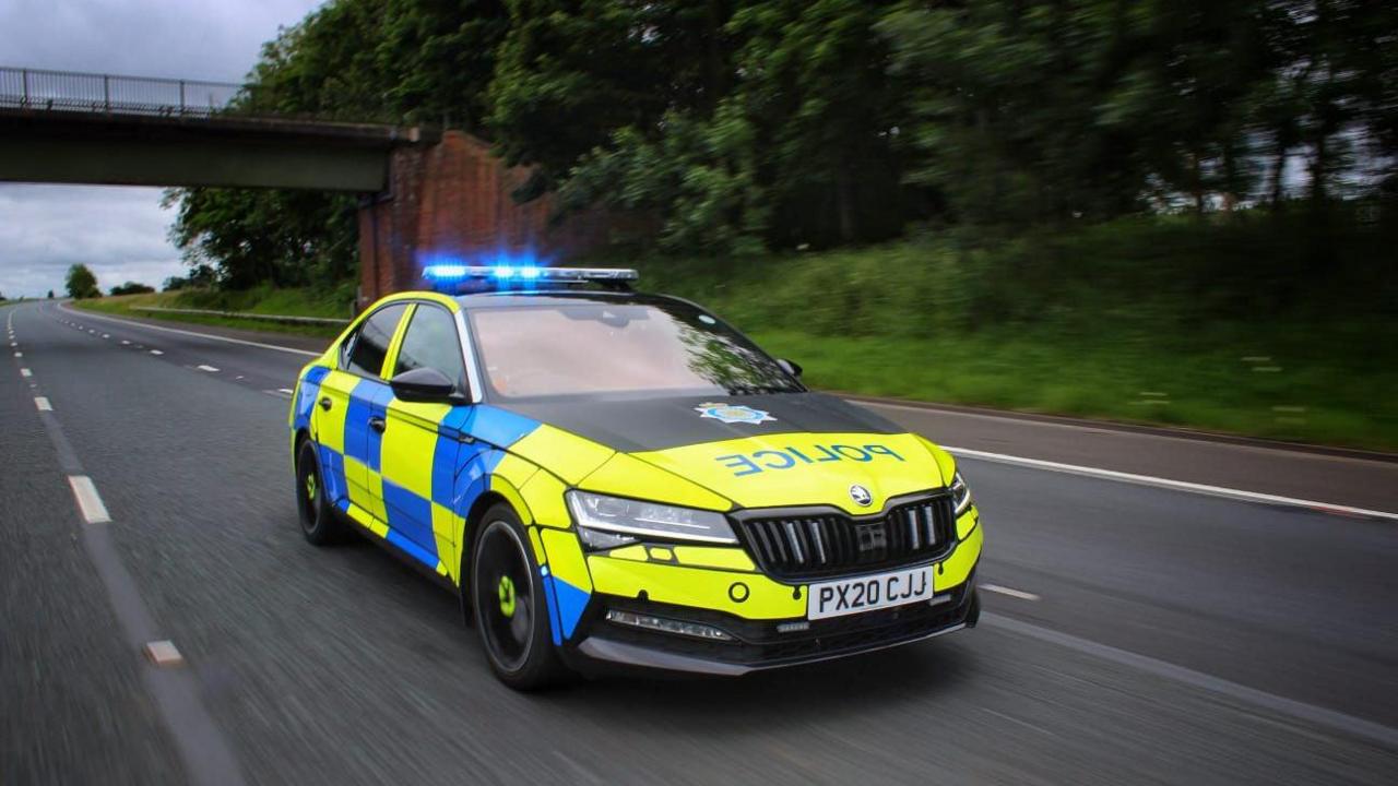 Stock image of a Cumbria Police car travelling along a road. The vehicle has a bright yellow and blue livery and its emergency lights are flashing.