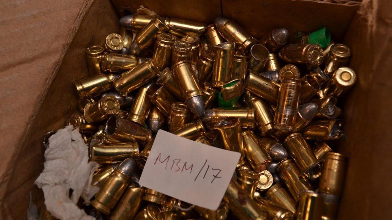 A cardboard box filled with gold-coloured bullet casings. A white card is lying in the box with "MBM/17" written on it. What appears to be a piece of tissue paper is also in the box.