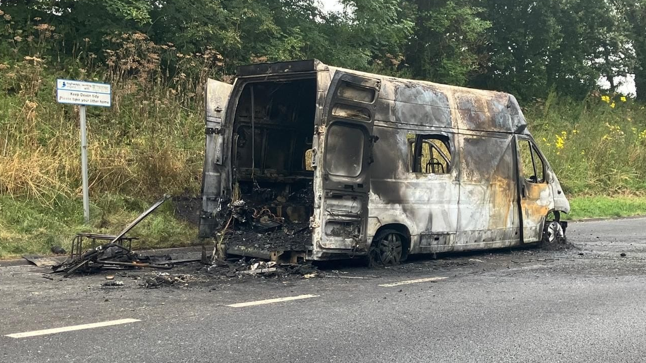 A burnt-out ambulance. The white paint is visible but all the markings have been burnt away, along with the tyres. There is what looks like ash in the rear of the ambulance and all over the road. 