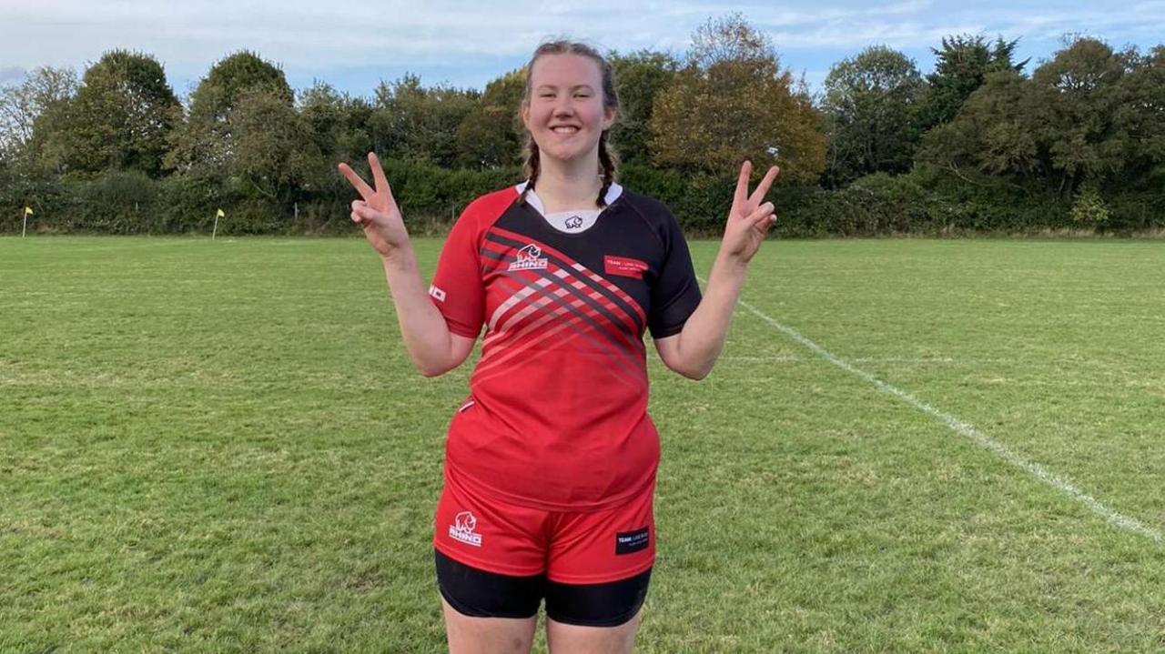 A young woman in a red sports top and shorts smiles proudly as she stands on a grass field. Her dark hair is in pigtails and she holds both hands up giving a v sign.