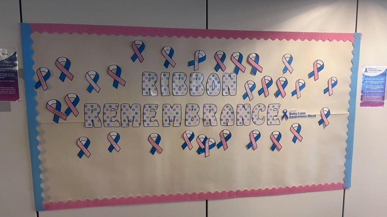 A display board with 'Ribbon Rememberance' written on it in a polka dot style. Dozens of pink and blue ribbons are placed around it.