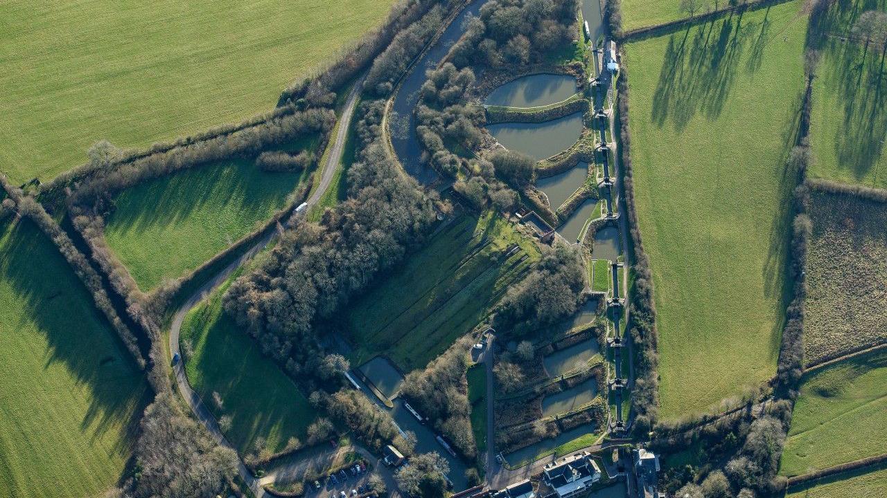 An aerial view of Foxton Locks in Leicestershire showing the staircase locks, surrounding by green fields and trees. A road can be seen to the left of the site.