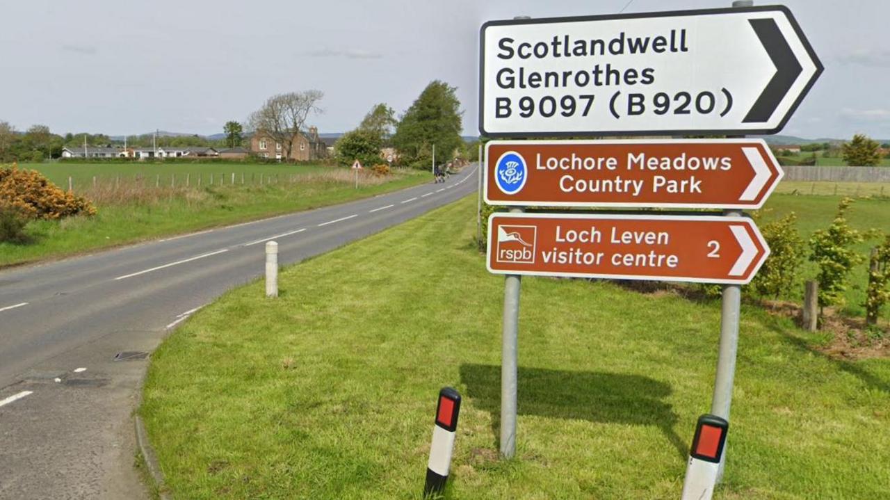 Picture of a road sign with a country road in the background. It features thee signs. One, which is white with black writing says "Scotlandwell Glenrothes B9097 (B920). Below this sign is two brown signs with white writing. One says: "Lochore Meadows Country Park" while the other says "Loch Leven visitor centre 2". All three signs are pointing the left. 