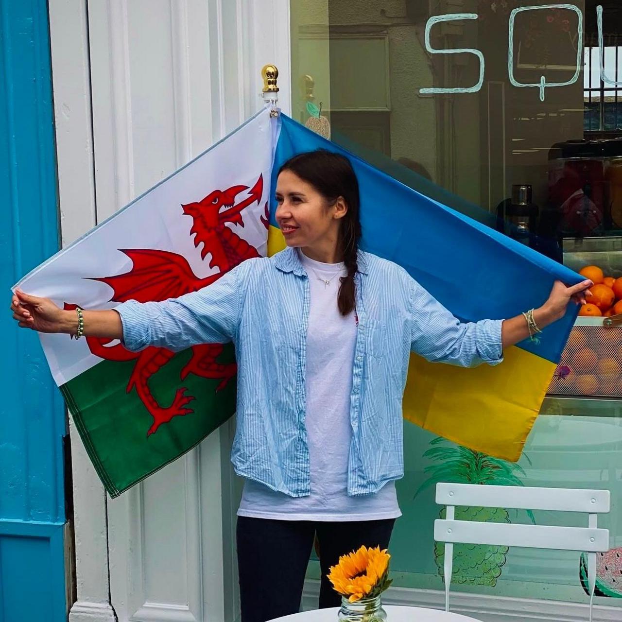 Maryna holding Wales and Ukraine flags. She is wearing a blue and white striped shirt with a white shirt underneath, and her long dark hair is in a plait. A box of oranges can be seen in the window in the background.