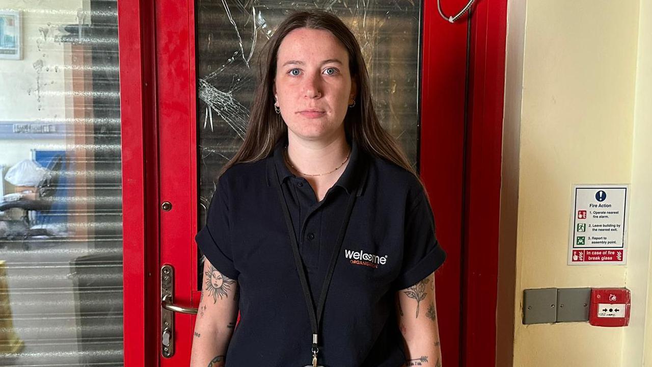 A woman with tattoos on her arms and long brown hair wearing a black polo shirt looks at the camera