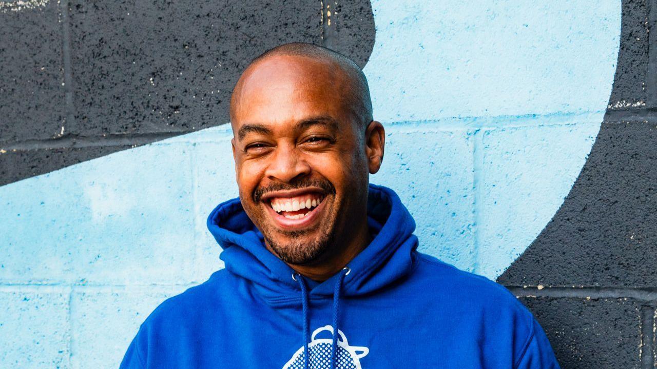 A man in a blue hoodie laughs as he stands in front of a grey wall with a blue stripe painted on it.