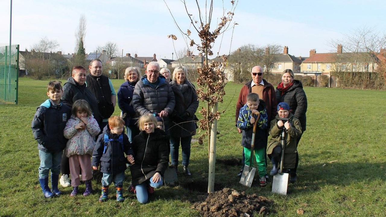 A number of adults and children standing in a park next to a newly planted tree 