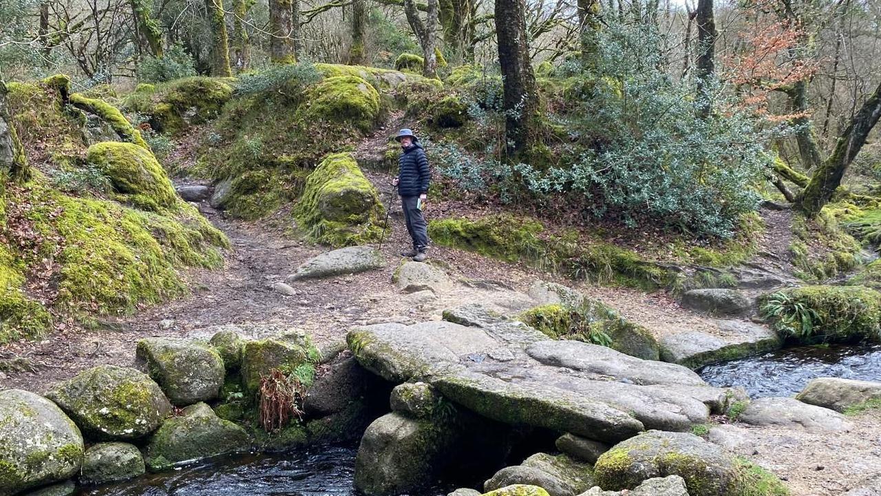 Gavin Robbins stands near a river on Dartmoor which has a stone-slabbed clapper bridge crossing it. He is wearing a large brimmed hat, a black coat, black trousers and has a walking pole in his right hand. In his left hand is a walking book.