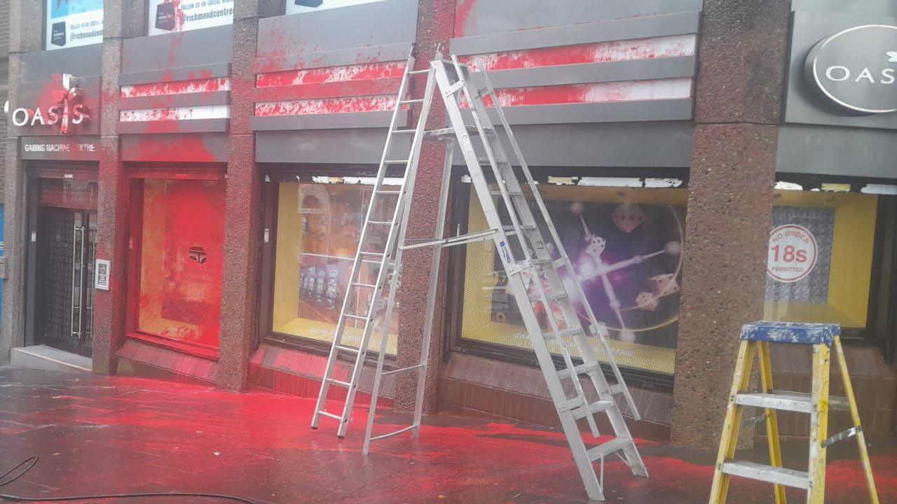 A shop front in Londonderry city centre partly covered in red paint following a vandalism attack. There are three large windows and a glass door all splattered with paint that is now partly cleaned. A ladder can be seen up against the building.