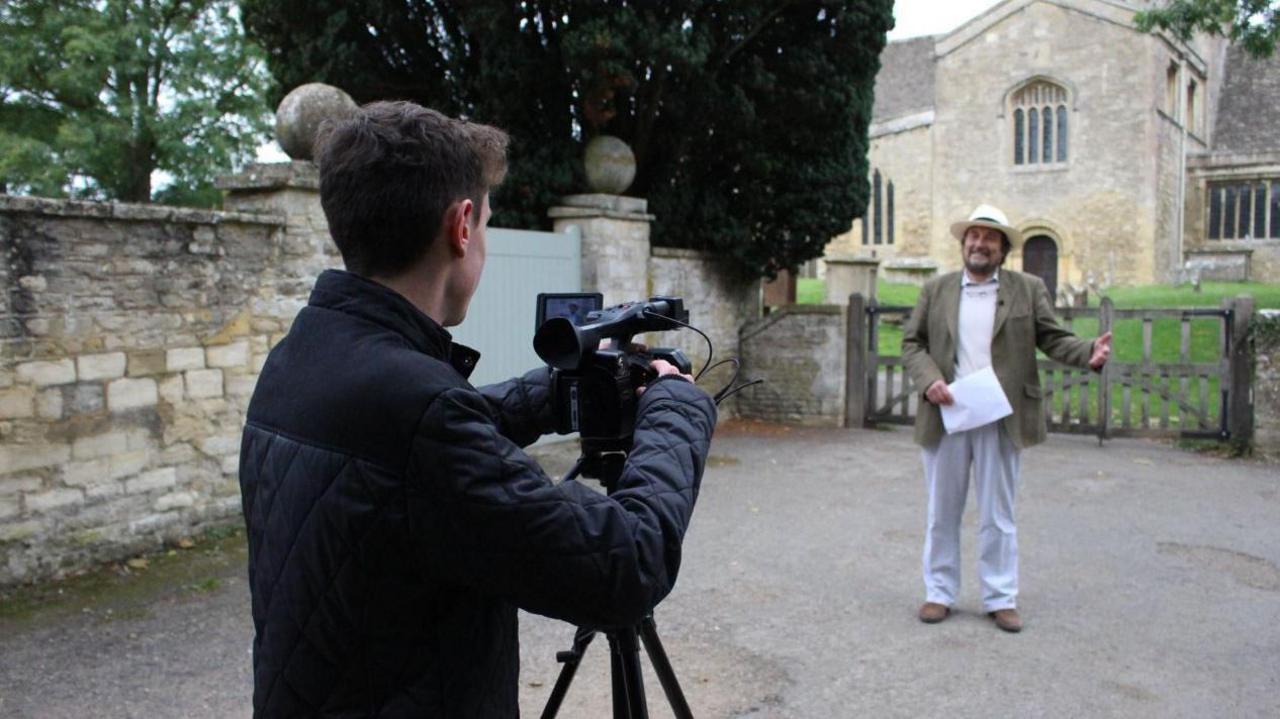 Robin Shuckburgh and his producer Ross Arrowsmith filming in front of a Cotswold church. Mr Shuckburgh is smiling while holding a piece of paper. Mr Arrowsmith is behind the camera filming him. It is a cloudy day.