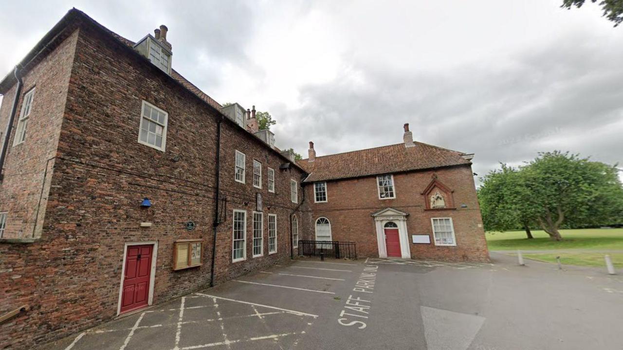 An L-shaped brick-built house, dating from the 1730s, has red doors and white window frames. It stands in a yard used for a car park, with a green field and trees in the background.