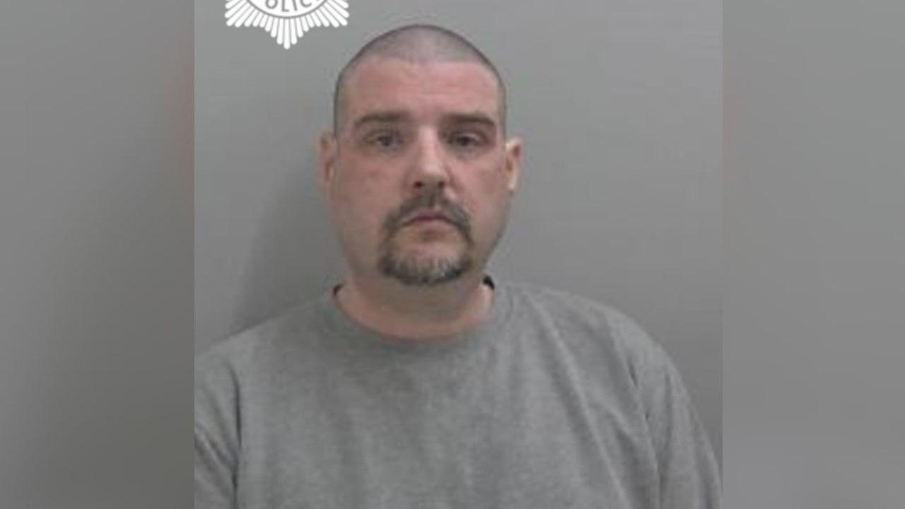 Chris Hemming pictured in a police mugshot. He has a blank expression and wears a grey jumper. He has a shaved head, dark stubbly hair and a dark beard.