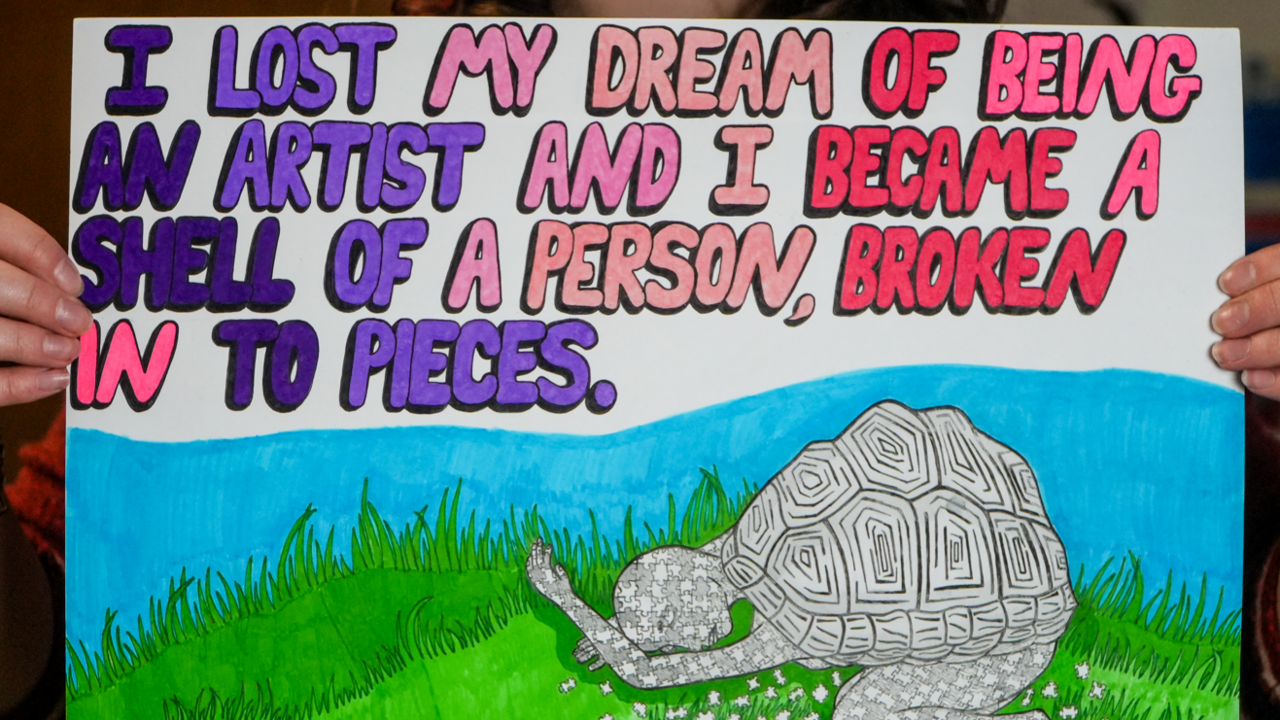 A poster depicts a person with a tortoise-like shell on their back while the are crouched in the grass. They lack on colour. Words on the poster read: "I lost my dream of being an artist and I became a shell of a person, broken into pieces."