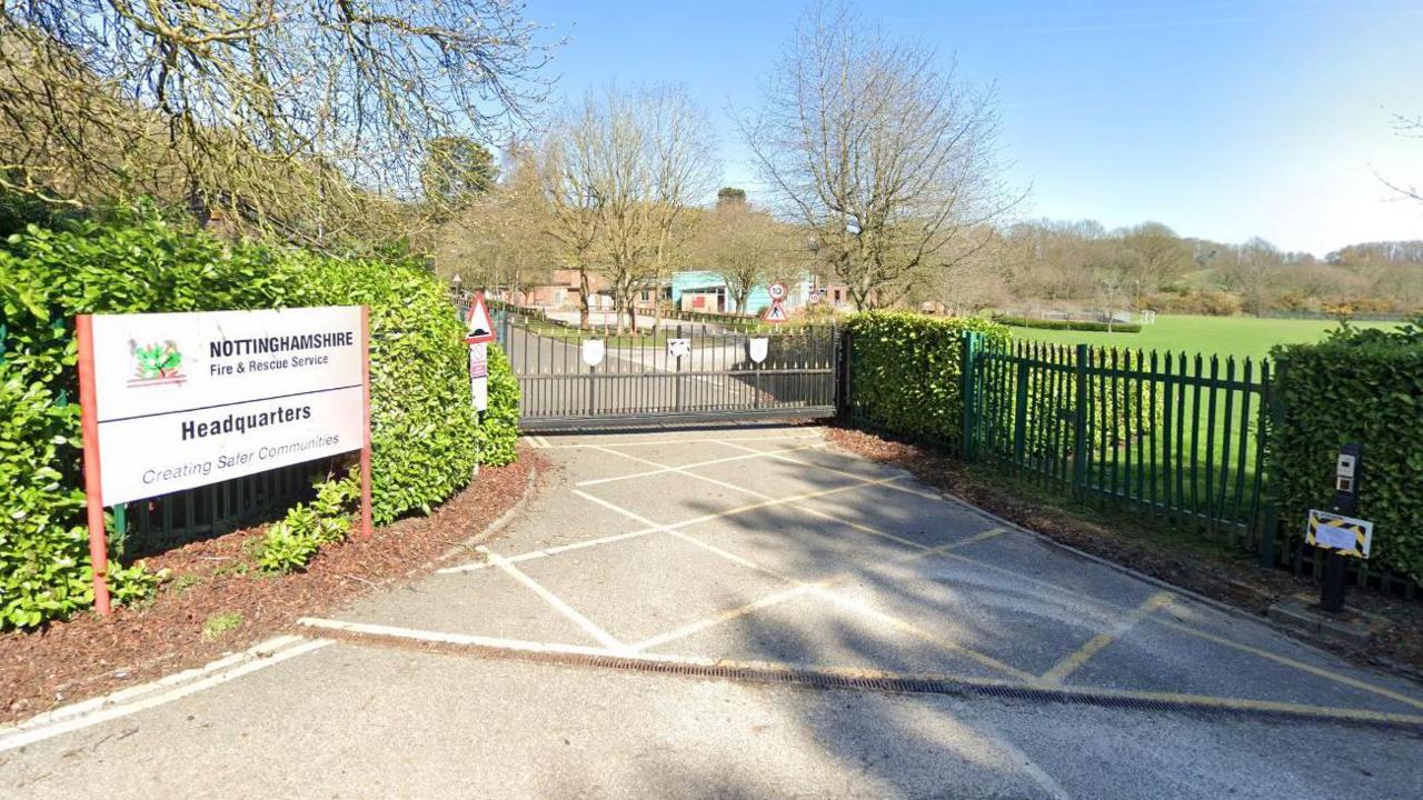 A Streetview image of the entrance to the old fire service headquarters, showing a gate and fence, with low brick buildings and an open field behind