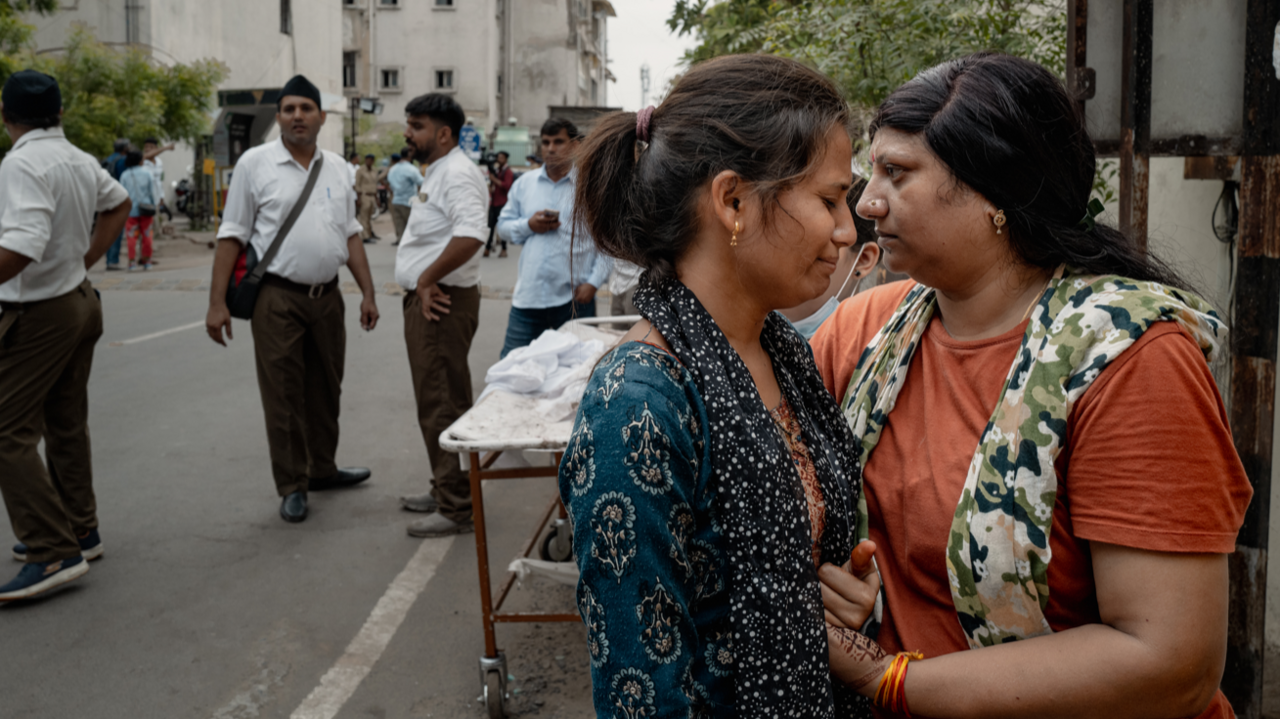 Two women, one wearing blue and one wearing orange, hold each other in the street. Men stand in the background, a trolley sits just behind them.