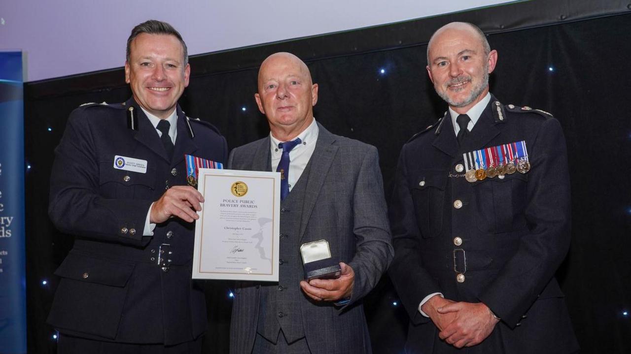 A man wearing a grey suit is receiving an award given to him by two police officers dressed in full uniform and medals