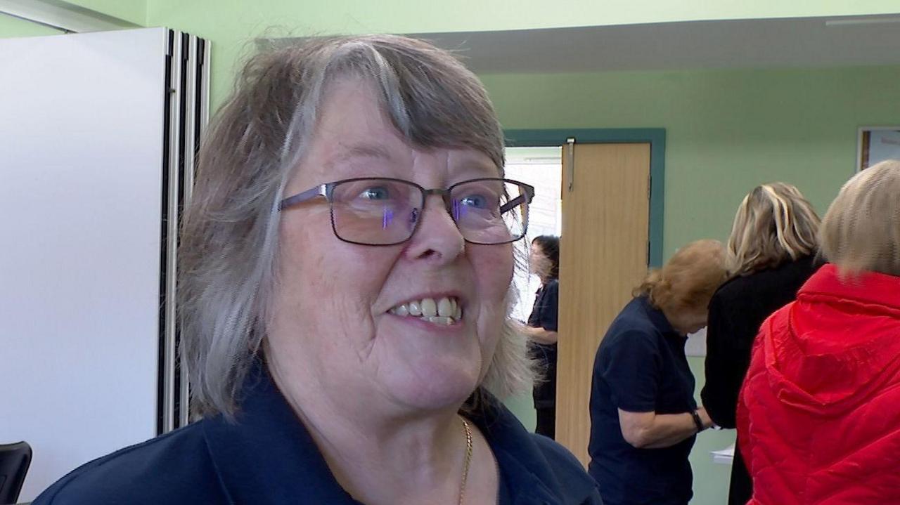 A woman stood in a room. She is wearing glasses and is wearing a blue polo shirt. She is smiling. There are people to the right of the woman in the background with their backs to the camera.