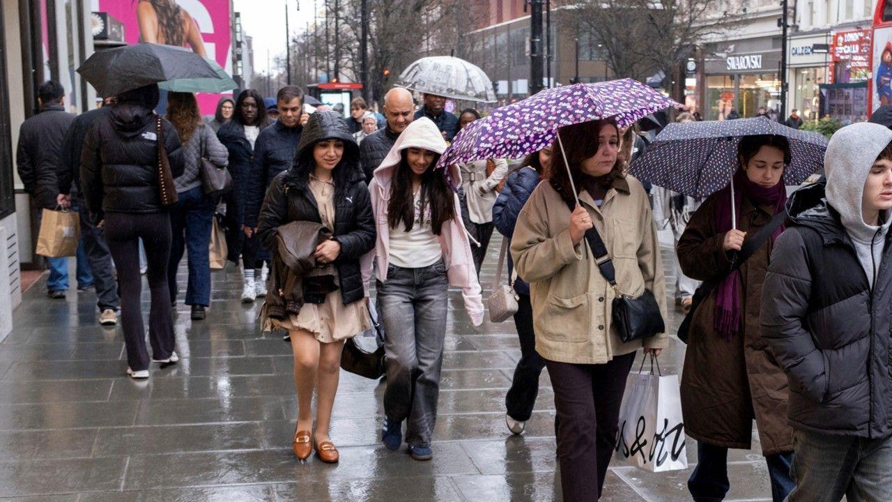 A crowd of people walk in both directions down a busy shopping street in the rain with hoods and umbrellas up