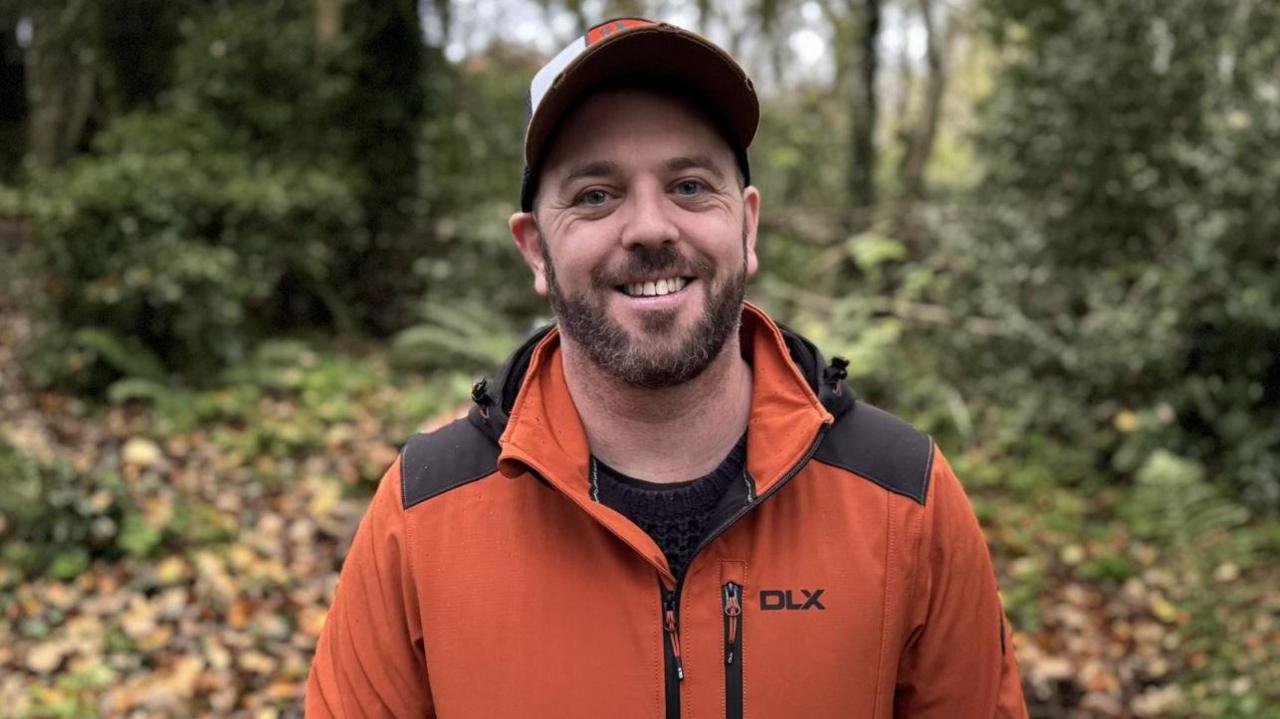 Keiron Brown, a man with a dark beard, an orange and white cap, a black top and an orange jacket with the words DLX on it.
