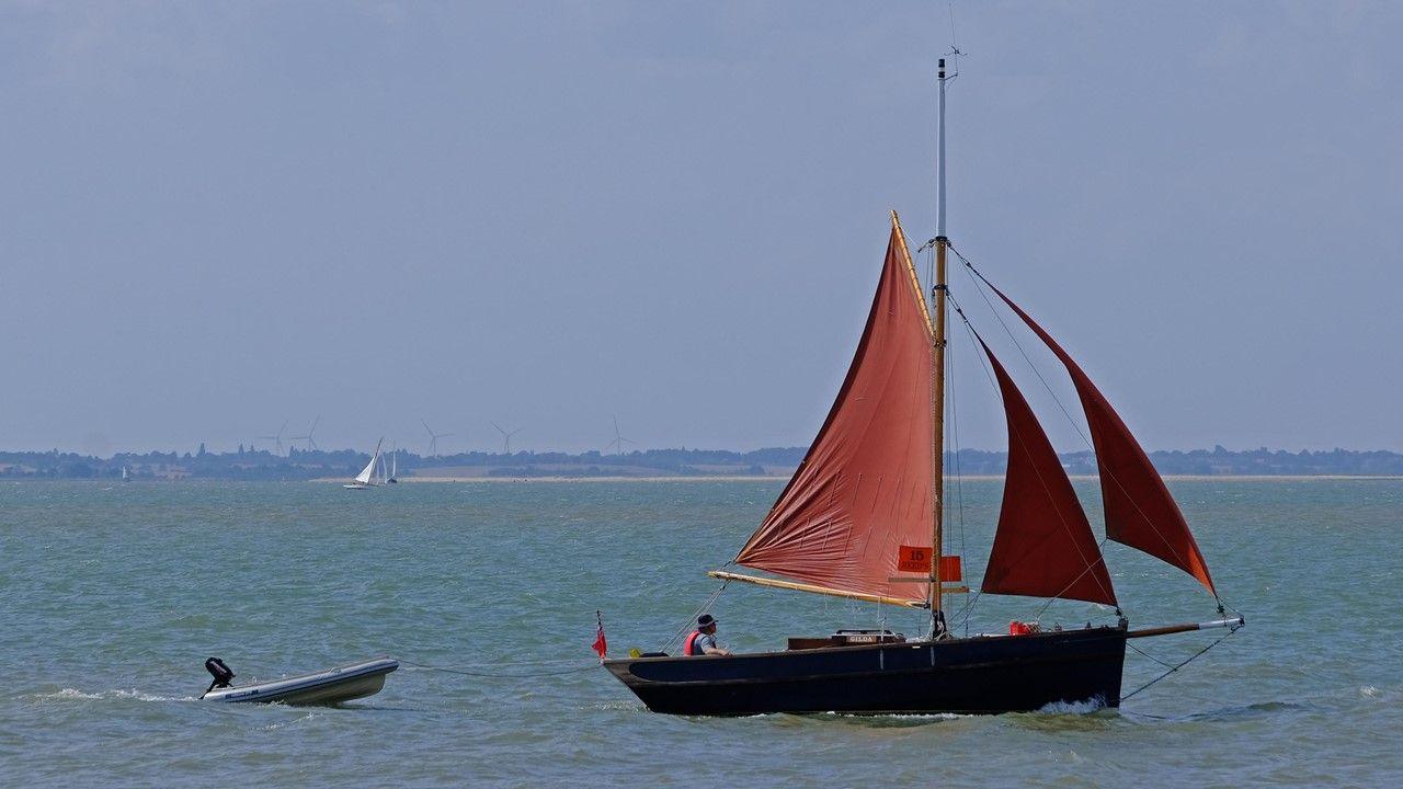 A boat with three open red sails draws a power boat behind it on the greeny blue sea with hazy blue sky above