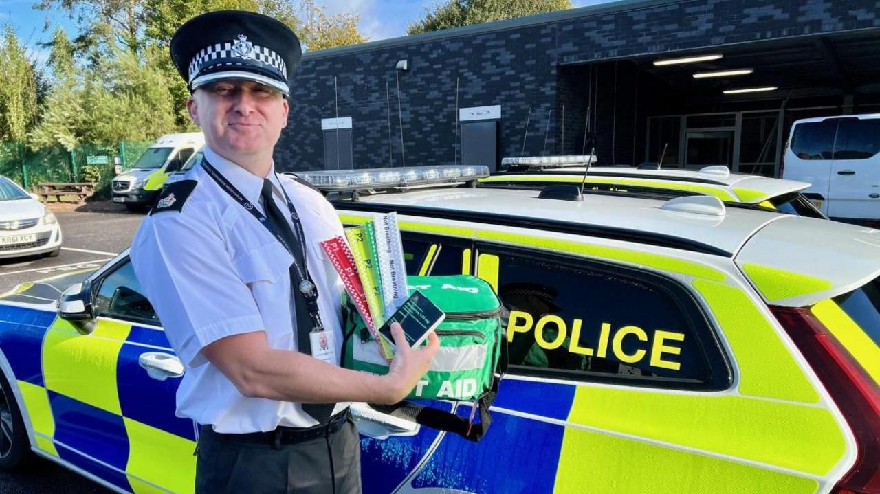 A police officer wearing a white short-sleeve shirt and a black police hat holding a green first aid kit bag while stood next to a blue and yellow police car in a car park.