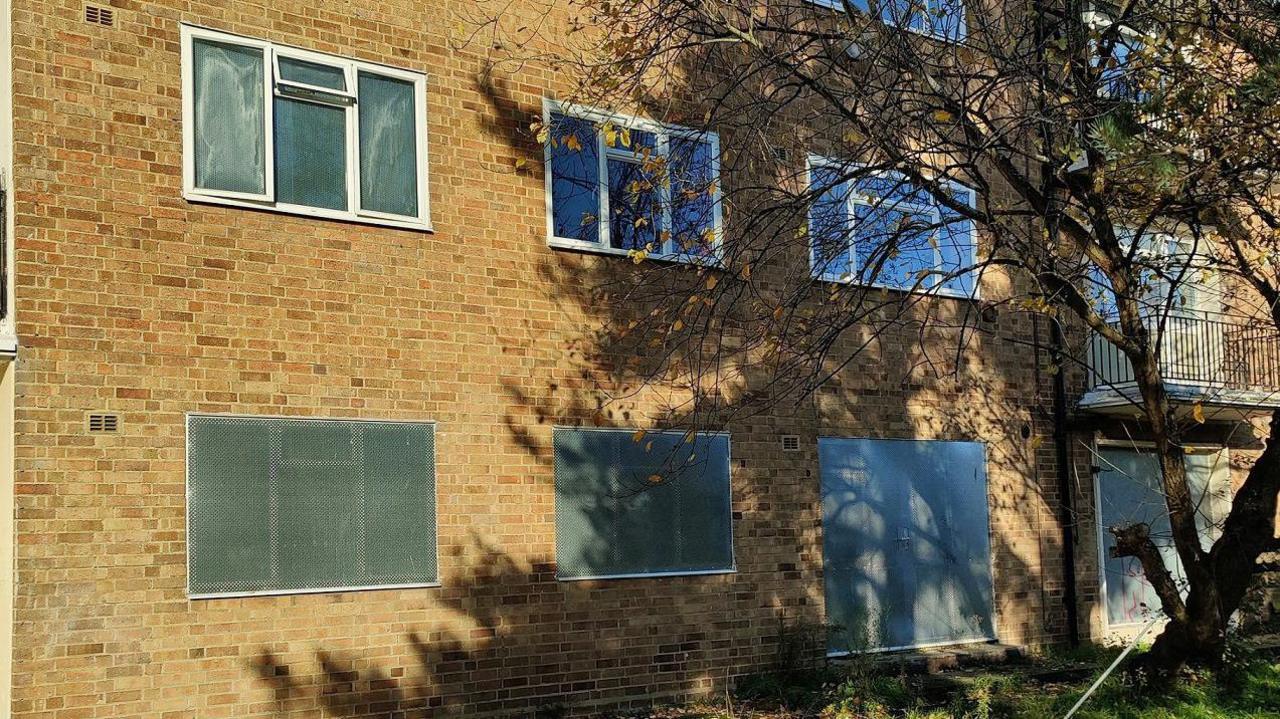 A block of flats with light brown bricks. There are nine windows visible. The three on the ground floor have been sealed with metal. There is a tree in front of the building on the right