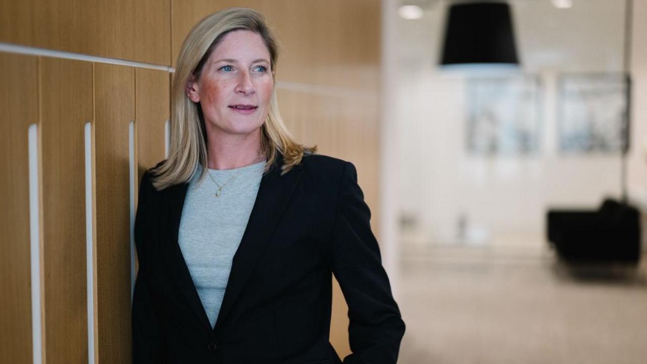 Sarah Emerson is a corporate photo. She has blonde hair and is wearing a slick black suit with a grey top and a necklace. We can see she is an office somewhere but the background is blurred.