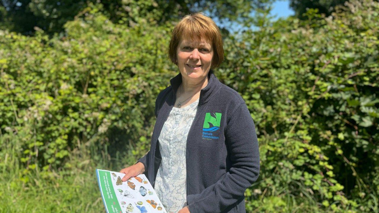 Angela Salmon standing outside, holding a chart of butterfly species