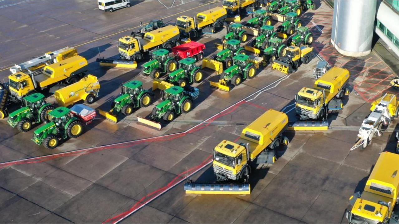 Green tractors and yellow HGV vehicles line up on the tarmac airside of Manchester Airport, each fitted with ploughs, brushes and other specialist equipment. 