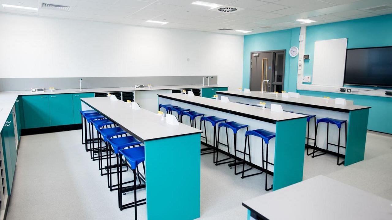 A room with three rows of tables with turquoise legs and metal-framed chairs arranged below them and more work surfaces around the walls
