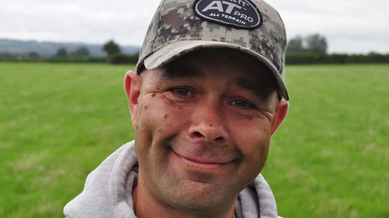 A closeup of a man with a grey cap and grey top in front of a green field