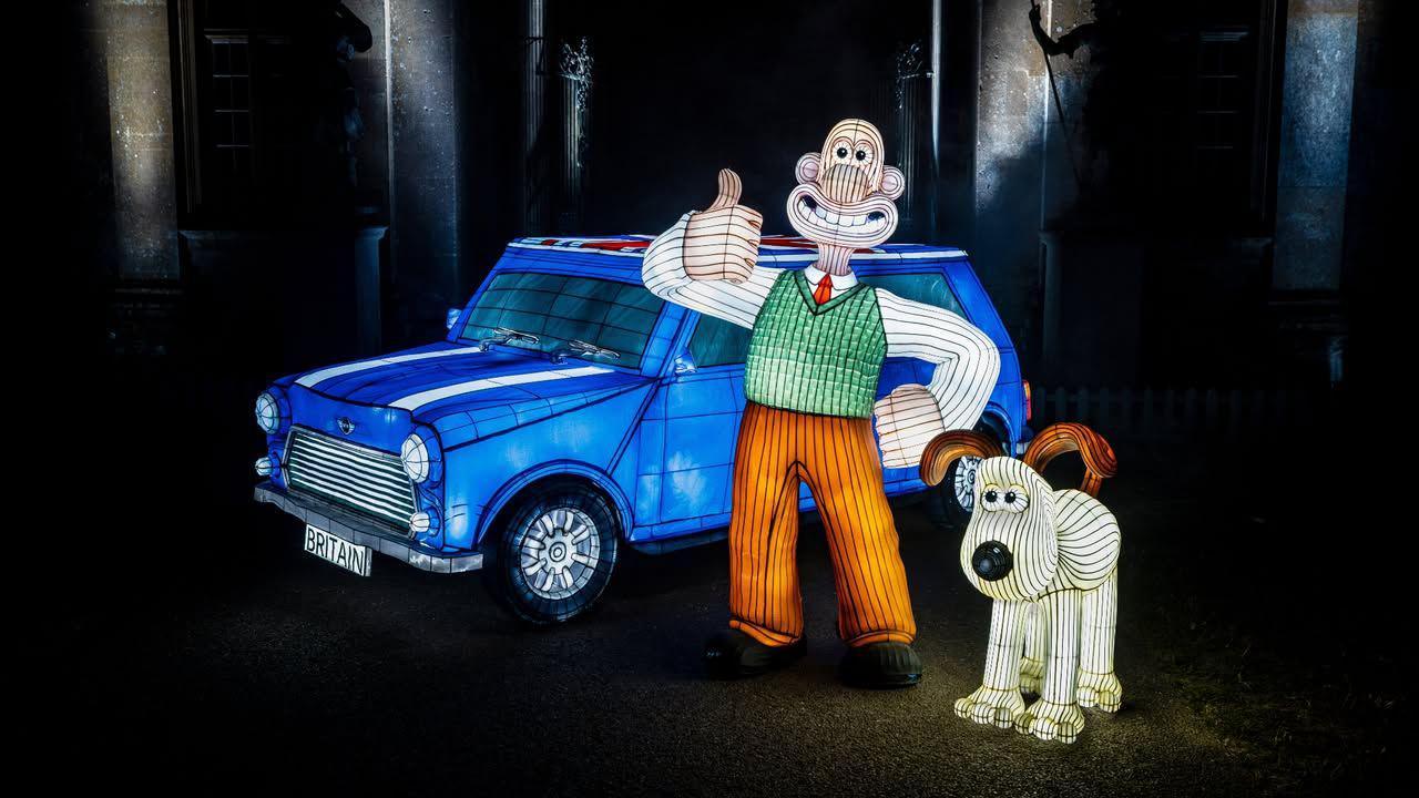 Three lanterns from Longleat's Festival of Light annual event. One is a blue car and the other two depict Wallace and Gromit - a stop-motion character wearing a white shirt, red tie, green jumper vest and orange trousers. He has one hand on his hip and a thumb up. Next to the Wallace lantern is Gromit, a cream dog with brown ears and a big black nose.