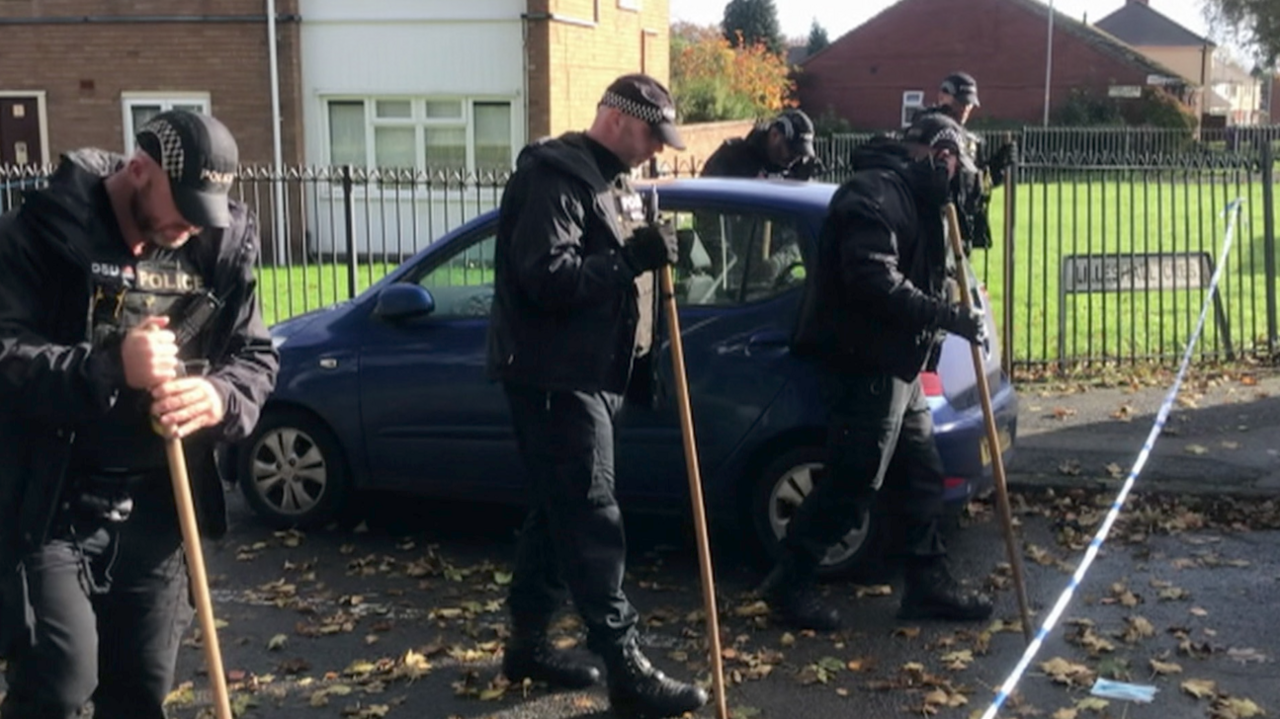 Police officers walk in a street scouring the ground with sticks by police tape. Three  stand in a line working together by a parked car close to houses.