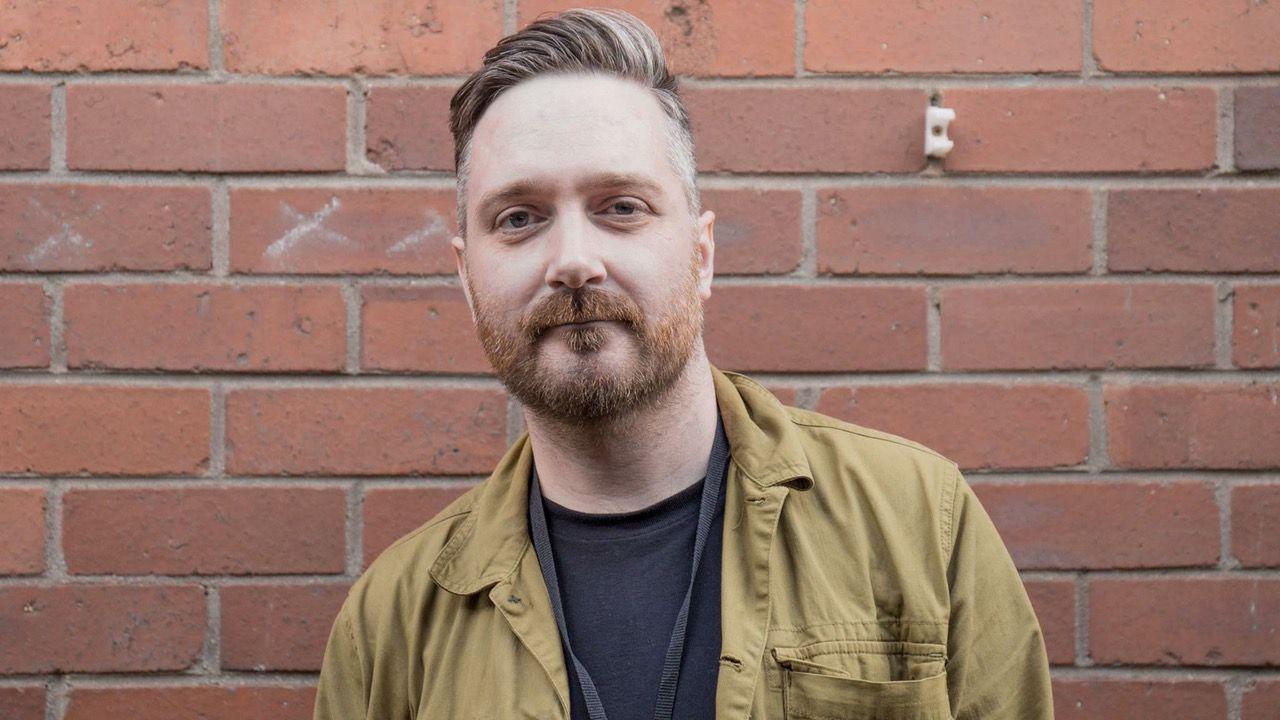 Photo of a man with a beard, green jacket against a red brick wall 