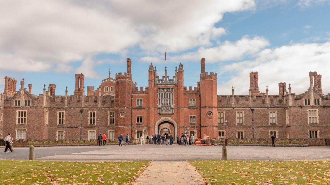 The front of a Tudor castle with an arched gateway entrance and a flag on top. Visitors and tourists are walking around a large courtyard at the front