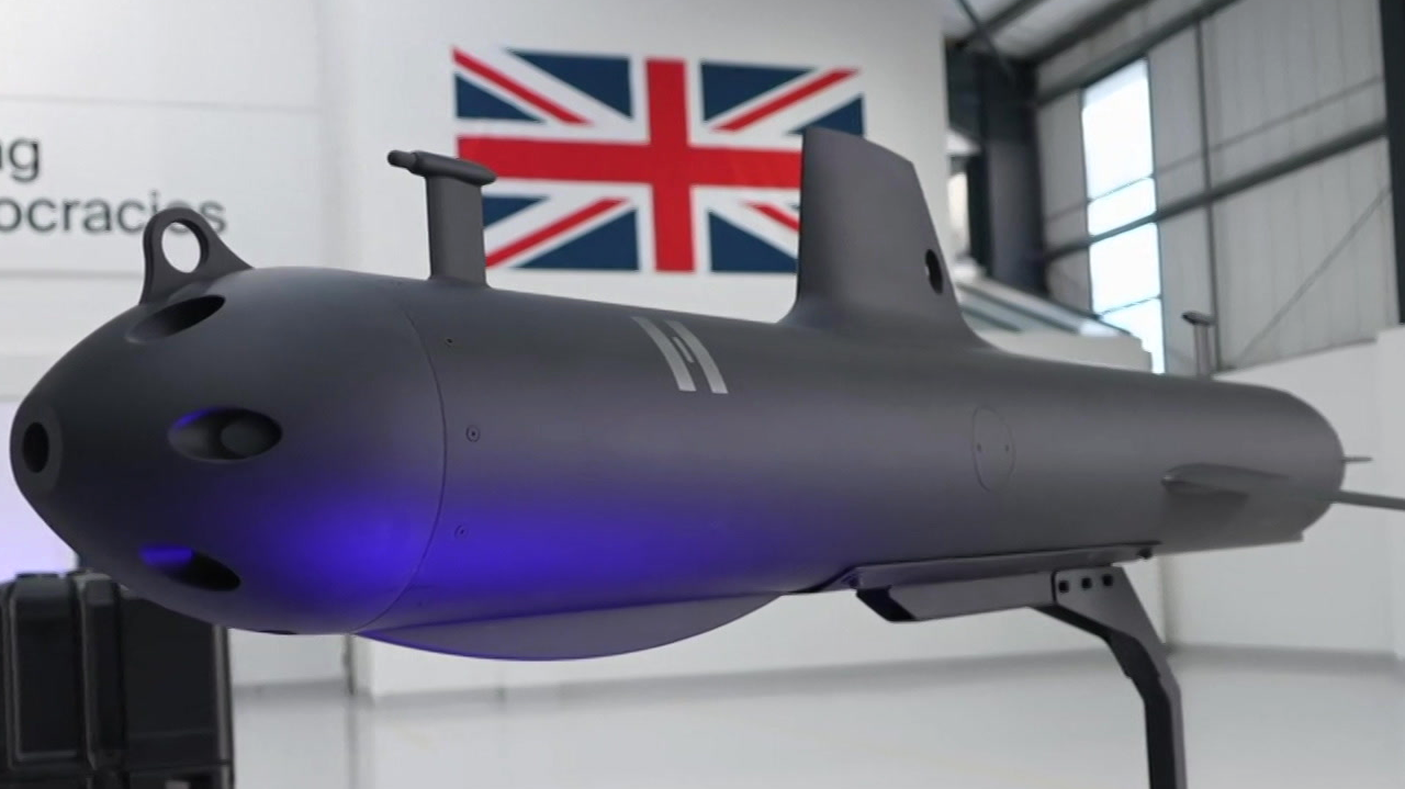 The image shows an unmanned submarine glider displayed indoors on a stand. The glider has a sleek, torpedo-like design with a matte dark finish and a pointed nose. It includes a vertical stabiliser fin and a long tail section for hydrodynamic control. A soft blue light is illuminating part of the body. In the background, there is a large Union Jack flag mounted on the wall and the setting appears to be a modern industrial or research space with white walls and high ceilings.