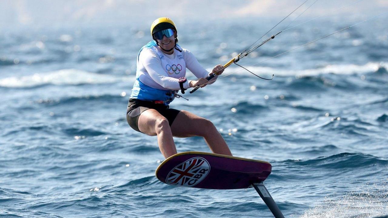 Ellie Aldridge smiling at the end of her kitesurf competition at the 2024 Olympics in Marseille. She is surrounded by water. Her feet are on the kiteboard and she is holding the bar and lines of her kite which is out of shot. She is wearing a yellow helmet and reflective goggles and a blue and white Team GB kit.