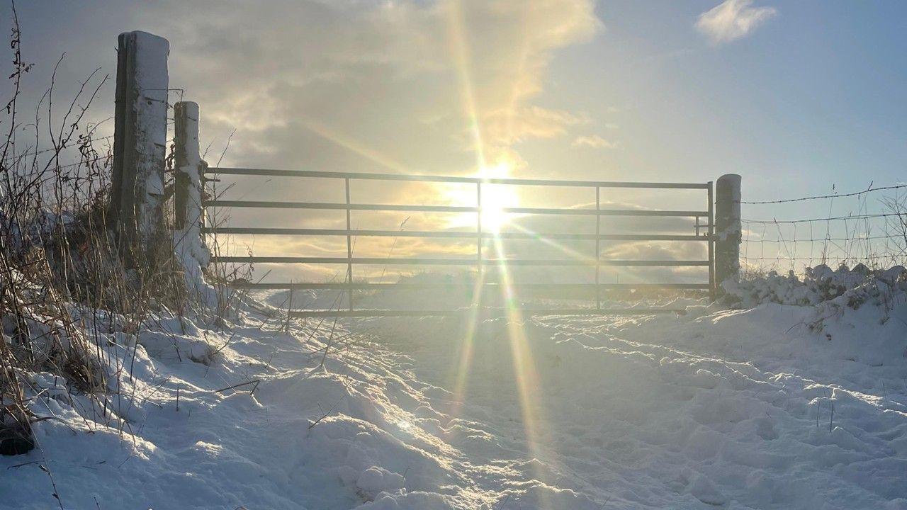A brilliant, diamond-like shot of sun rays coming through a wide country gate with snow on the ground in front and yellow and blue colours in the sky