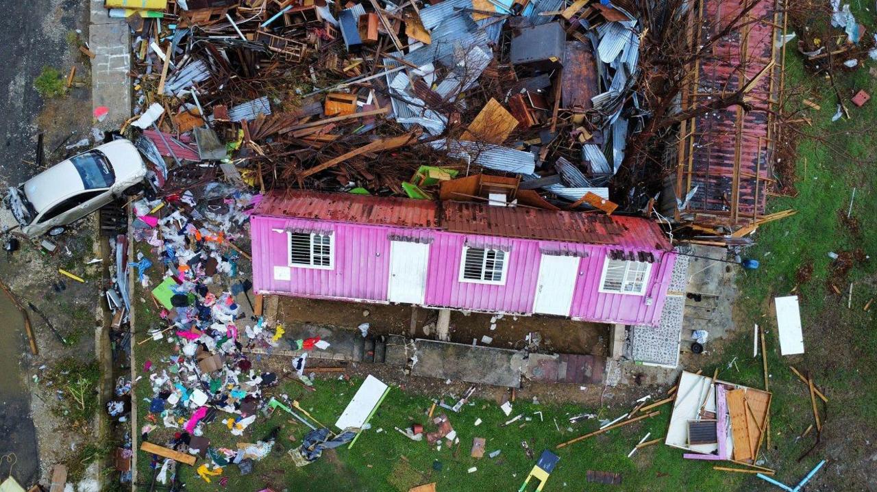 An overhead drone view showing a pink one-storey corrugated metal home, which has been turned over on its back by the hurricane. It had three windos and two white doors. Debris completely surrounds the structure with a damaged silver car on the left.