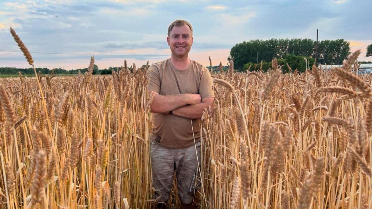 Chris Dodson smiles at the camera as he stands in a field of straw with his arms crossed across his chest. He is wearing a brown T-shirt and jeans. Behind him is a blue and pink skyline. 